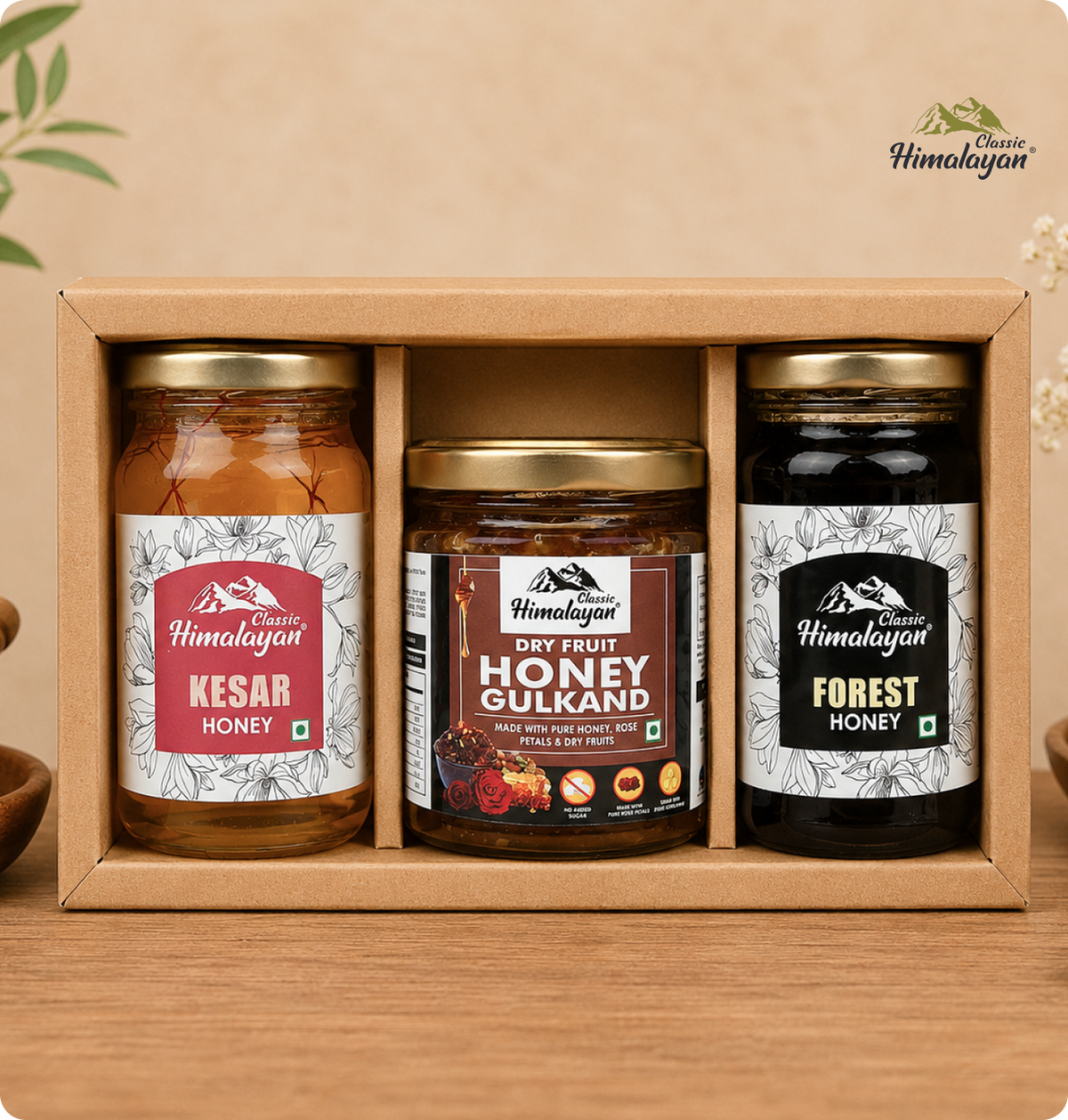 A Himalayan gift set containing three jars of honey in a light brown cardboard box: Kesar Honey, Dry Fruit Honey Gulkand, and Forest Honey, displayed on a wooden surface against a warm beige background.