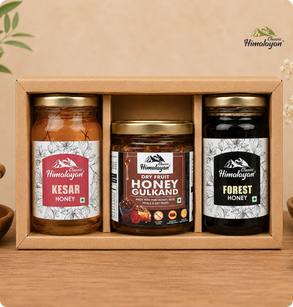 A Himalayan gift set containing three jars of honey in a light brown cardboard box: Kesar Honey, Dry Fruit Honey Gulkand, and Forest Honey, displayed on a wooden surface against a warm beige background.